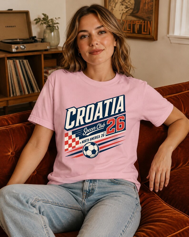 K&ouml;nnte beinhalten: Ein hellrosa T-Shirt mit kurzen &Auml;rmeln und einem Grafikdruck mit der Aufschrift CROATIA Soccer Club 26 North America 26. Das Design enth&auml;lt ein rot-wei&szlig; kariertes Quadrat, einen Fu&szlig;ball und blaue sowie rote horizontale Linien.