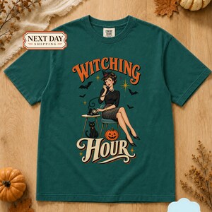 May include: An emerald green short-sleeve t-shirt featuring a graphic print of a person in a witch hat sitting on a chair, talking on a vintage telephone. The design includes a black cat, a jack-o'-lantern, bats, and the text Witching Hour.