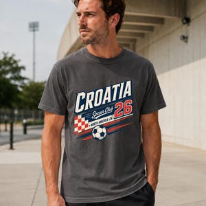 K&ouml;nnte beinhalten: Ein anthrazitfarbenes Kurzarm-T-Shirt mit einem Grafikdruck, der CROATIA Soccer Club 26 NORTH AMERICA 26 zeigt. Das Design enth&auml;lt ein rot-wei&szlig;es Karomuster, einen Fu&szlig;ball sowie blaue und rote horizontale Bewegungslinien.