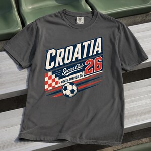 K&ouml;nnte beinhalten: Ein anthrazitfarbenes T-Shirt mit einem Grafikdesign, das den Text CROATIA, Soccer Club, 26 und NORTH AMERICA 26 enth&auml;lt. Das Design zeigt ein rot-wei&szlig;es Karomuster, einen Fu&szlig;ball sowie blaue und rote horizontale Bewegungslinien.