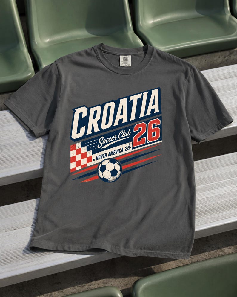 K&ouml;nnte beinhalten: Ein anthrazitfarbenes T-Shirt mit einem Grafikdesign, das den Text CROATIA, Soccer Club, 26 und NORTH AMERICA 26 enth&auml;lt. Das Design zeigt ein rot-wei&szlig;es Karomuster, einen Fu&szlig;ball sowie blaue und rote horizontale Bewegungslinien.