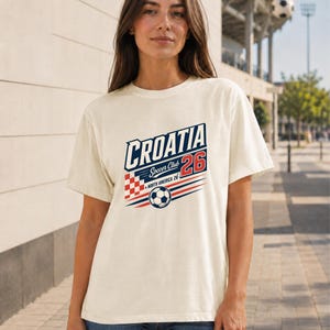 K&ouml;nnte beinhalten: Ein cremefarbenes Kurzarm-T-Shirt mit einem Grafikdesign, das den Text CROATIA, Soccer Club und NORTH AMERICA 26 enth&auml;lt. Das Design zeigt ein rot-wei&szlig;es Karomuster, einen Fu&szlig;ball sowie rote und blaue horizontale Linien.