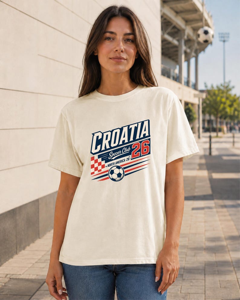 K&ouml;nnte beinhalten: Ein cremefarbenes Kurzarm-T-Shirt mit einem Grafikdesign, das den Text CROATIA, Soccer Club und NORTH AMERICA 26 enth&auml;lt. Das Design zeigt ein rot-wei&szlig;es Karomuster, einen Fu&szlig;ball sowie rote und blaue horizontale Linien.