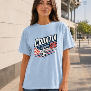 K&ouml;nnte beinhalten: Ein hellblaues Kurzarm-T-Shirt mit einem Grafikdruck, der CROATIA Soccer Club 26 North America 26 zeigt. Das Design enth&auml;lt ein rot-wei&szlig;es Karomuster, einen Fu&szlig;ball sowie rote und blaue horizontale Bewegungslinien.