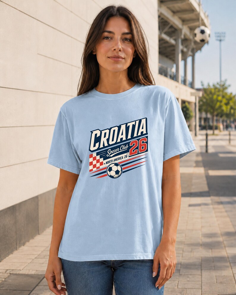 K&ouml;nnte beinhalten: Ein hellblaues Kurzarm-T-Shirt mit einem Grafikdruck, der CROATIA Soccer Club 26 North America 26 zeigt. Das Design enth&auml;lt ein rot-wei&szlig;es Karomuster, einen Fu&szlig;ball sowie rote und blaue horizontale Bewegungslinien.