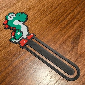 May include: A black bookmark with a green and red pixelated Yoshi character from the Super Mario Bros. video game series.