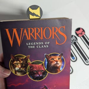 May include: A set of four book marks featuring different cat designs. The book marks are made of black plastic with a yellow, pink, or blue accent. The book marks are on a white background. The book marks are for the book "Warriors: Legends of the Clans".