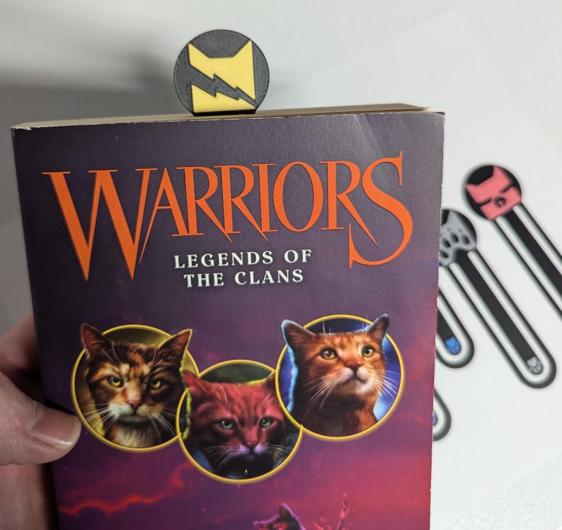 May include: A set of four book marks featuring different cat designs. The book marks are made of black plastic with a yellow, pink, or blue accent. The book marks are on a white background. The book marks are for the book "Warriors: Legends of the Clans".