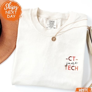 May include: Cream-colored t-shirt with the text "-CT- jane TECH" in pink and black. A gold necklace with a pendant rests on the shirt. The shirt is folded and next to a brown hat.