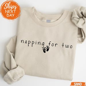 May include: A sand-colored sweatshirt with the phrase "napping for two" in black script, accompanied by two black footprint graphics. The sweatshirt has a crew neck and long sleeves, perfect for casual wear.