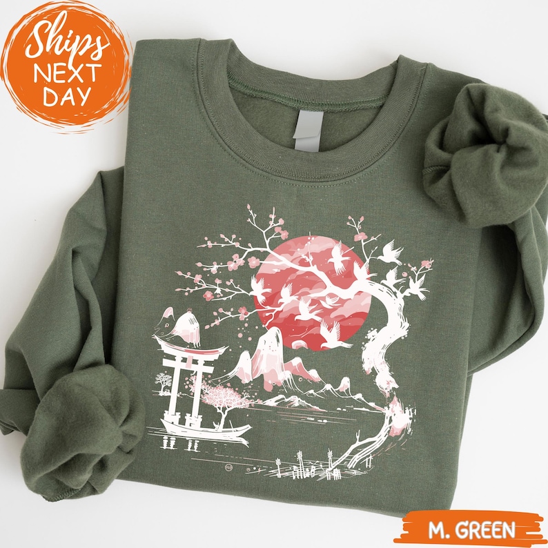 A green sweatshirt with a graphic design depicting a cherry blossom tree with a red moon and mountains in the background.