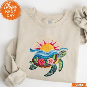May include: Sand-colored sweatshirt featuring an embroidered design of a sea turtle with a sunset and ocean waves. The turtle has a green head and flippers, a brown shell with pink hibiscus flowers, and a blue and green patterned design. The text "Ships Next Day" is in the upper left corner.