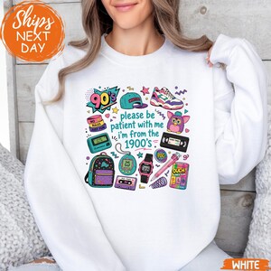 May include: A white crewneck sweatshirt featuring a colorful graphic print of 1990s nostalgia items, including a pager, cassette tape, digital watch, and a Furby. The text reads, "90's please be patient with me i'm from the 1900's" in a playful font.