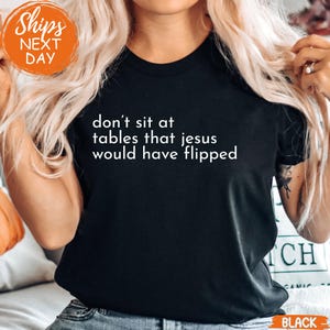 May include: Black t-shirt with the white text "don't sit at tables that jesus would have flipped." The shirt has a crew neck and short sleeves. The image also includes an orange "Ships Next Day" graphic.