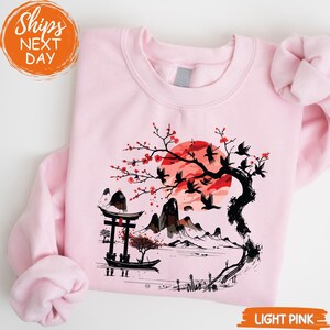 A pink sweatshirt with a graphic design depicting a cherry blossom tree with red flowers and a temple, along with mountains and a sunset in the background.