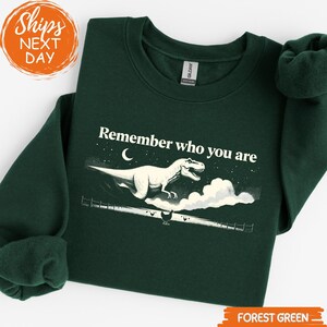 May include: Forest green sweatshirt with the text "Remember who you are" and a white dinosaur graphic. The design includes a crescent moon, stars, and clouds. The sweatshirt is a Gildan brand.