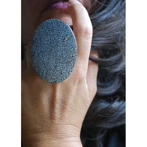 May include: A large oval ring featuring a textured gray and black surface with a mottled, organic pattern. The ring has a smooth metallic border and is worn on a finger, showcasing its substantial size and detailed, intricate surface design.