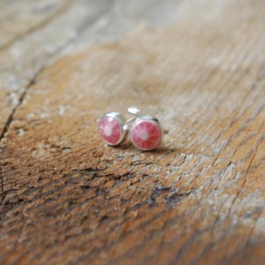 May include: A pair of round silver cufflinks with a pink and white floral design. The cufflinks are set against a rustic wooden background, showcasing their delicate details and contrasting textures.
