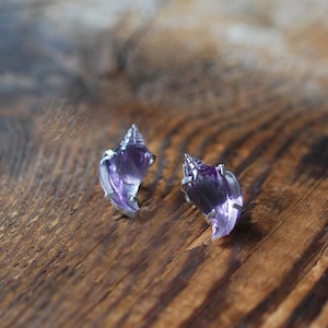 May include: A pair of stud earrings featuring carved purple amethyst gemstones shaped like small spiral seashells. Each translucent purple stone is secured in a simple silver-toned metal prong setting, highlighting the natural color variations.