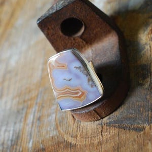 Pode incluir: Um anel com uma pedra polida em forma de escudo, apresentando faixas concêntricas nas cores lavanda, branco e castanho. A pedra está engastada numa moldura de prata lisa, exibindo camadas minerais naturais e uma inclusão cristalina num dos lados.