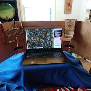 Puede incluir: Un ordenador portátil con un marco de madera y una manta azul sobre la mesa. La pantalla del portátil muestra un mapa con un esquema de color verde y marrón. Hay una torre de dados de madera en el lado derecho del portátil.