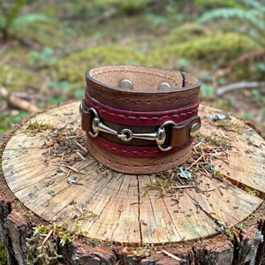 May include: A wide leather cuff bracelet featuring a central silver-toned metal horse bit hardware piece. The bracelet is constructed from layered brown and magenta leather strips with visible stitching and two silver-toned metal snap closures.