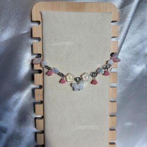 Può includere: Un bracciale a catena color argento con un ciondolo centrale a forma di farfalla bianca, intervallato da piccole perline a forma di fiore rosa e bianco, perline a forma di stella e ciondoli rosa traslucidi a forma di campanella. Il testo Lovertrinkets è visibile.