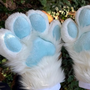 May include: A pair of white and light blue paw gloves. The gloves have a fluffy white exterior with light blue paw pads. The wrist cuffs are white. The gloves are designed to resemble animal paws.