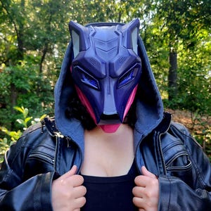 May include: A dark purple and pink wolf mask with pointed ears and angular geometric features. The mask covers the upper face and is worn with a black hooded jacket over a black top. The design features sharp lines and a stylised animal aesthetic.