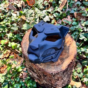 May include: A matte black, stylised feline mask featuring geometric, angular panels and recessed eye openings. The mask has a structured, robotic appearance with textured ventilation details on the snout and forehead, crafted in a solid, dark finish.