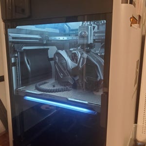 May include: A Bambu Lab H2S 3D printer with a white exterior and a black front frame. The glass door reveals an internal print bed containing a black, complex geometric object and a long, segmented black print component under blue lighting.