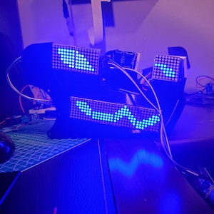Op de afbeelding: Een zwart elektronisch apparaat met drie afzonderlijke LED-puntmatrixpanelen die heldere blauwe lichtpatronen weergeven. De panelen zijn verbonden door blootliggende bedrading en gemonteerd op een donker frame, wat een modulair digitaal displaysysteem vormt.