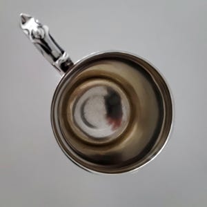 Puede incluir: Una taza de plata con un asa decorativa. La taza est&aacute; vac&iacute;a y tiene una superficie brillante y reflectante.