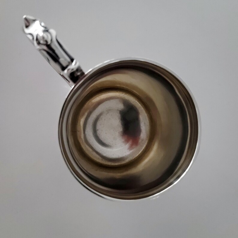 Puede incluir: Una taza de plata con un asa decorativa. La taza est&aacute; vac&iacute;a y tiene una superficie brillante y reflectante.
