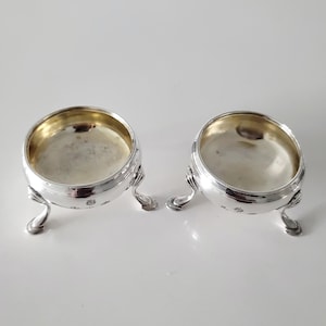May include: Two antique silver salt cellars. Each small bowl has a rounded shape, a shiny interior, and rests on three curved legs. The rims are detailed with a decorative pattern. The salt cellars are displayed on a white surface.