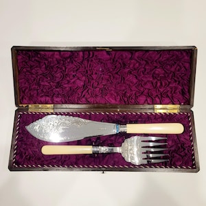 Puede incluir: Un juego de servir de plata vintage, que incluye un cuchillo y un tenedor, presentado en una caja de madera con un forro de terciopelo morado. La hoja del cuchillo presenta un diseño floral ornamentado.
