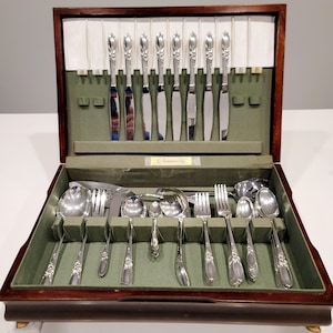 Puede incluir: Un cofre de cubiertos de madera con un interior de fieltro verde, que contiene un juego completo de cubiertos de color plateado. El juego incluye cuchillos, tenedores, cucharas y utensilios para servir. El cofre tiene una etiqueta "Community".