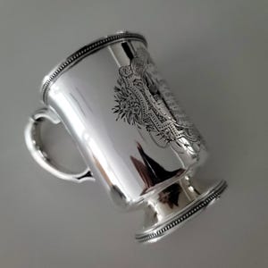 Puede incluir: Taza de plata con un dise&ntilde;o grabado y un asa. La taza tiene una base redonda y un borde con cuentas.