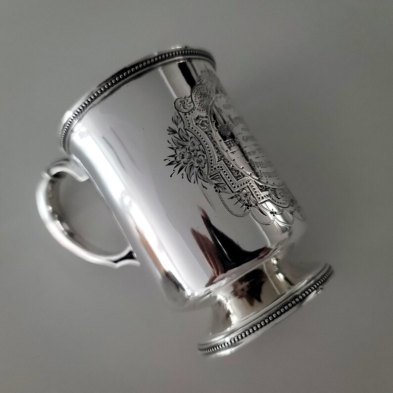 Puede incluir: Taza de plata con un dise&ntilde;o grabado y un asa. La taza tiene una base redonda y un borde con cuentas.