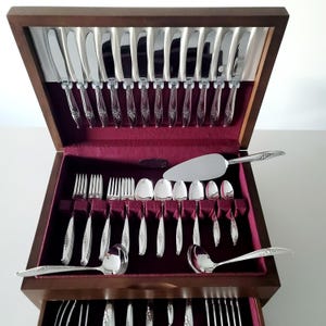 Puede incluir: Un cofre de cubiertos de madera con un interior de terciopelo burdeos, lleno de un juego completo de cubiertos plateados. El juego incluye cuchillos, tenedores, cucharas y utensilios para servir, todos cuidadosamente organizados.