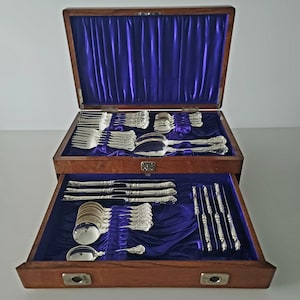May include: A wooden cutlery chest with a blue velvet lining, containing a full set of ornate silver utensils. The set includes forks, spoons, knives, and serving pieces, all neatly arranged within the chest.