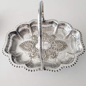 May include: A silver-colored, ornate serving tray with a handle. The tray has a scalloped edge and is decorated with floral and scrollwork patterns. The handle is also silver and has a textured design. The tray is likely made of metal and is designed for serving food or displaying items.