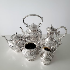 Op de afbeelding: Een zilveren theeservies met sierlijke details, waaronder een theepot, een melkkan, een suikerpot en twee andere vaten. De set heeft ingewikkelde patronen en gebogen handvatten. De stukken zijn op een wit oppervlak geplaatst.