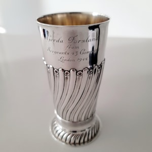 Op de afbeelding: Een zilverkleurige vintage beker met een uitlopende bovenkant en een decoratief, wervelend patroon op de body. De beker heeft een gegraveerde inscriptie: "Gerda Forsland from Sergeants 43 Company London 1944."