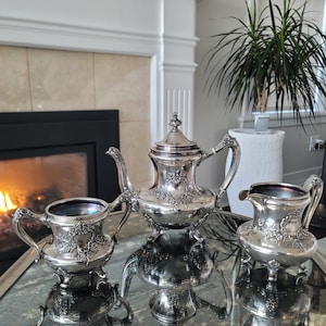 Puede incluir: Un juego de té de plata con tetera, jarra de crema y azucarero. El juego presenta un diseño floral ornamentado y está en excelente estado.