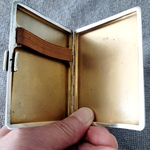 May include: Open silver cigarette case with a gold-colored interior. The case has a brown elastic band and a clasp closure. The interior is smooth and reflective, with a small hallmark stamp visible.