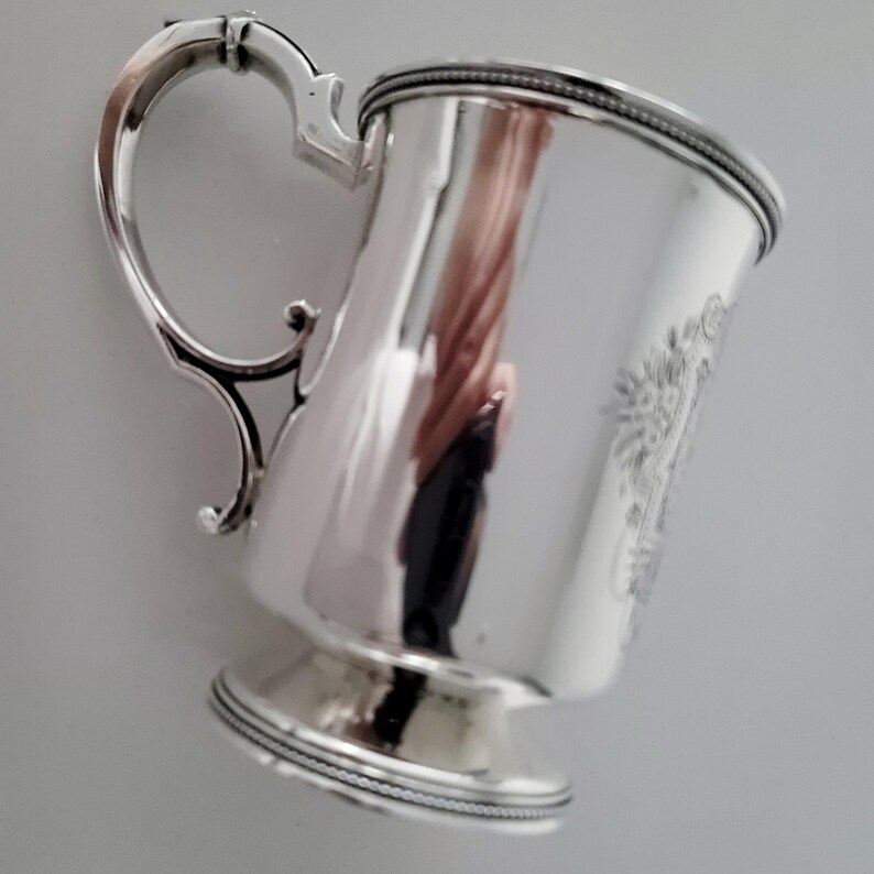 Puede incluir: Una taza de plata con un asa decorativa y un borde de cuentas. La taza tiene una base redondeada y una superficie lisa y pulida.