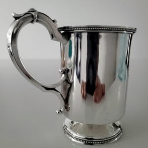 Puede incluir: Una taza de plata con un asa decorativa y un borde con cuentas. La taza tiene una base redonda y est&aacute; pulida.