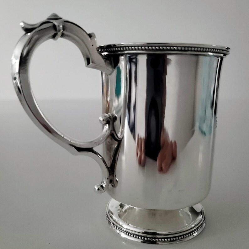 Puede incluir: Una taza de plata con un asa decorativa y un borde con cuentas. La taza tiene una base redonda y est&aacute; pulida.