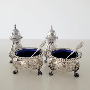 Puede incluir: Un juego de saleros y pimenteros de plata antiguos. Los saleros tienen un interior azul y están sostenidos por patas de león. Los pimenteros tienen un diseño calado decorativo. Cada salero tiene una cuchara pequeña.